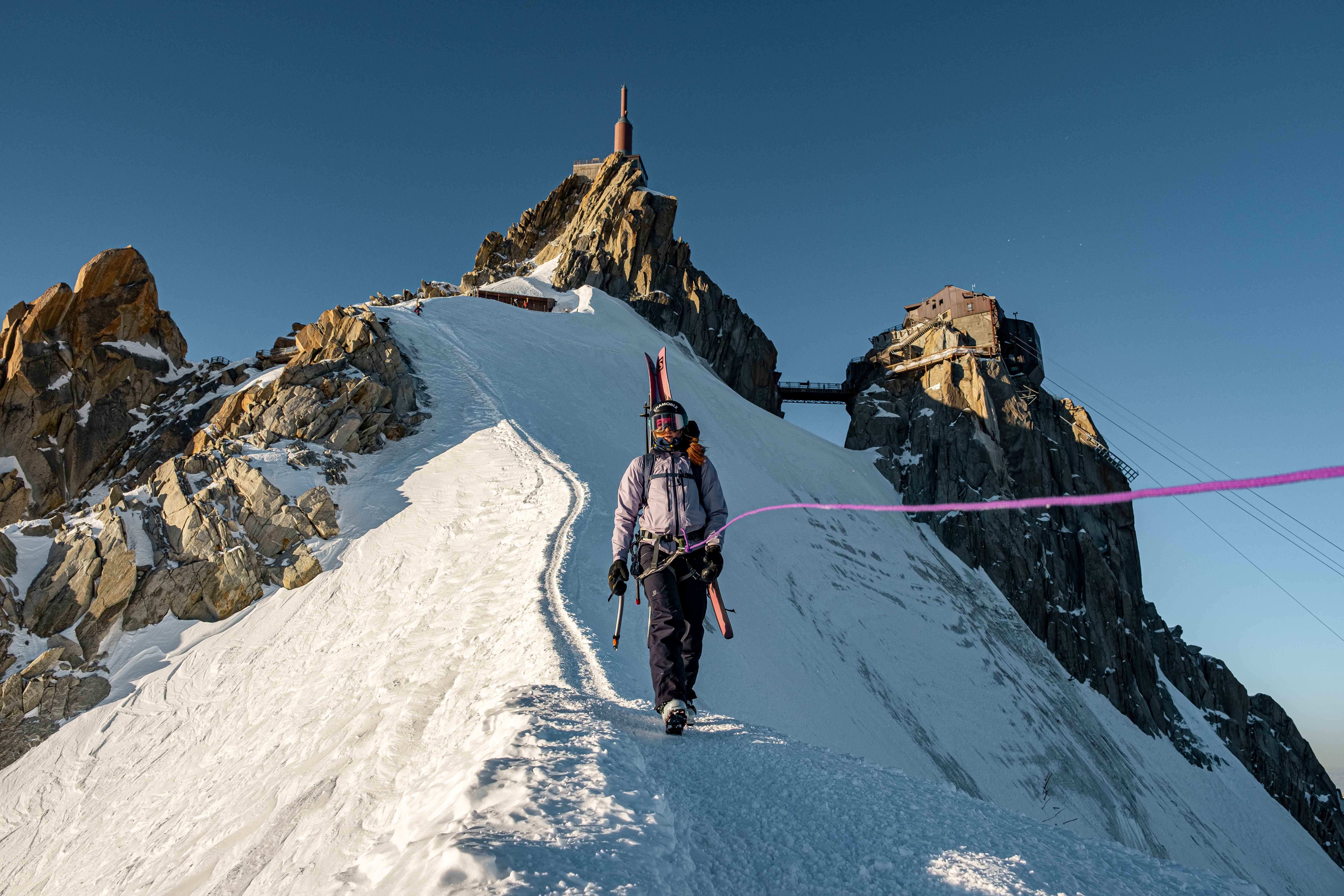 Woman in a skisuit walks across a mountain ridge attached to a rope on top of the Aiguille de Midi