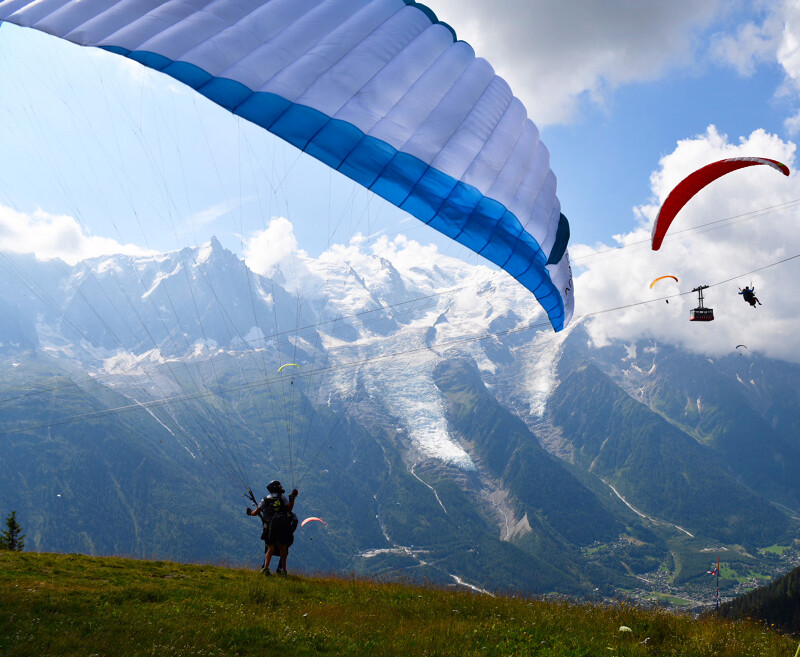 Two people about to take off on a green mountain with a paraglide, overlooking the mountains and cable cars of the valley 