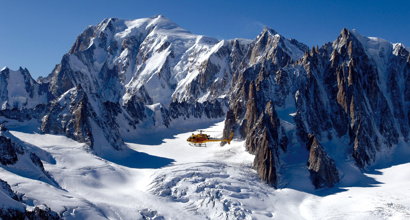 yellow helicopter hovering above a mountain chain on a sunny day